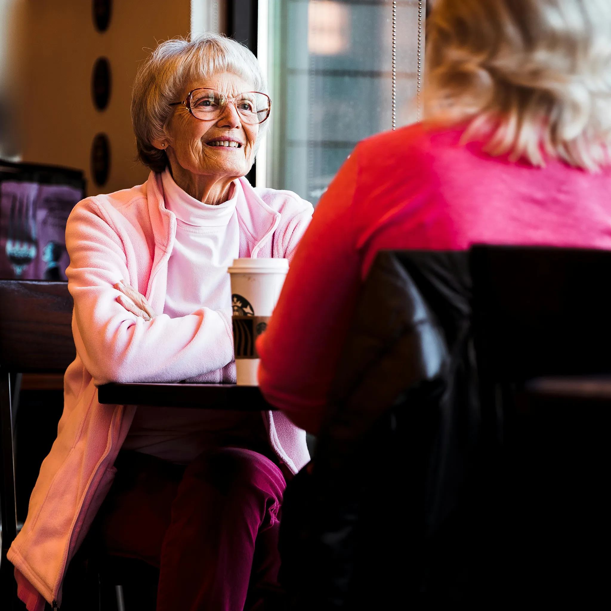 An elderly woman with short gray hair and glasses smiles while looking right, wearing a pink fleece jacket. She is seated at a dark table with a Starbucks coffee cup.