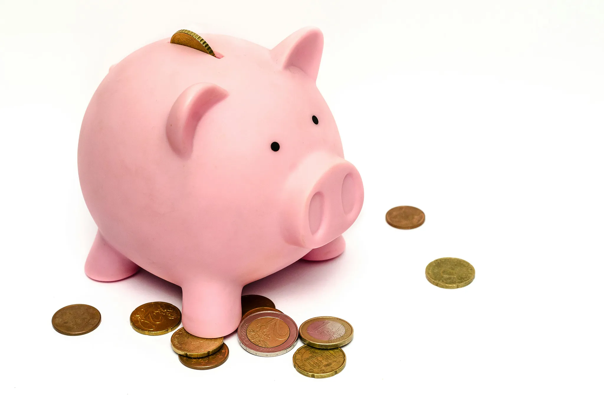 A pink piggy bank sits on a white surface, with a coin inserted in its back and several other coins scattered around it.
