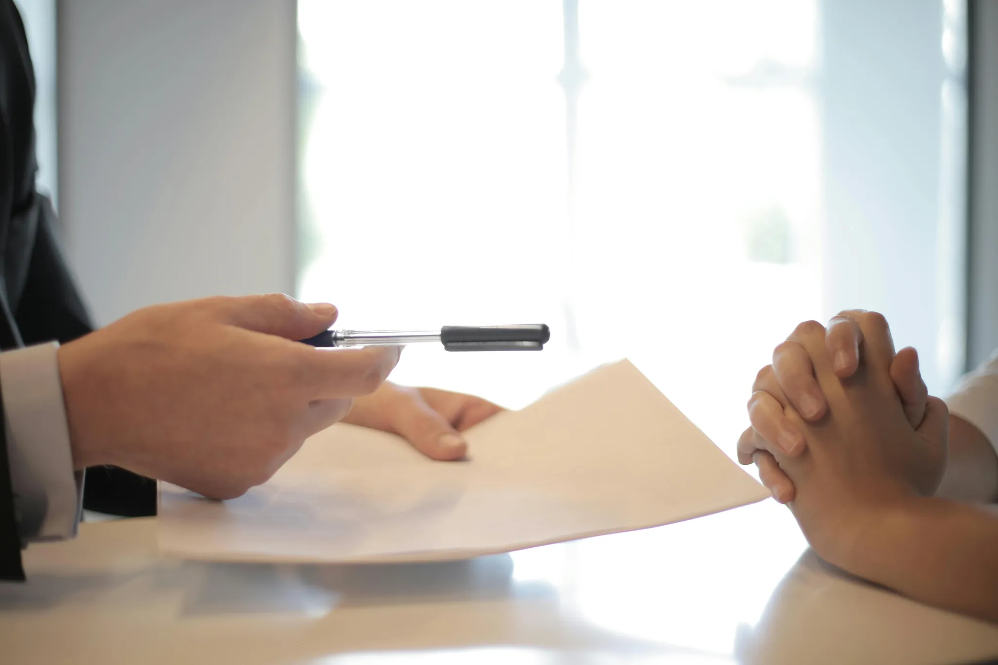 A person in a suit holds a pen over a document while another person's clasped hands rest on the table.