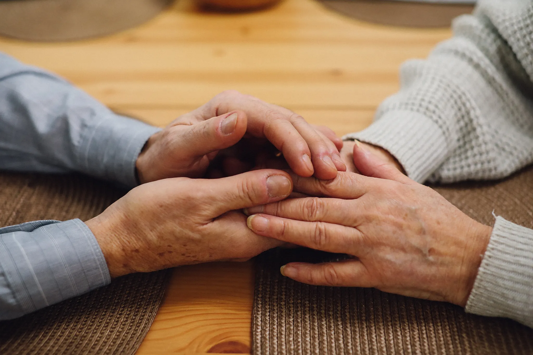 Two sets of hands, one male and one female, are gently clasped together on a woven placemat. The male wears a light blue striped shirt, and the female wears a light gray knitted sweater.