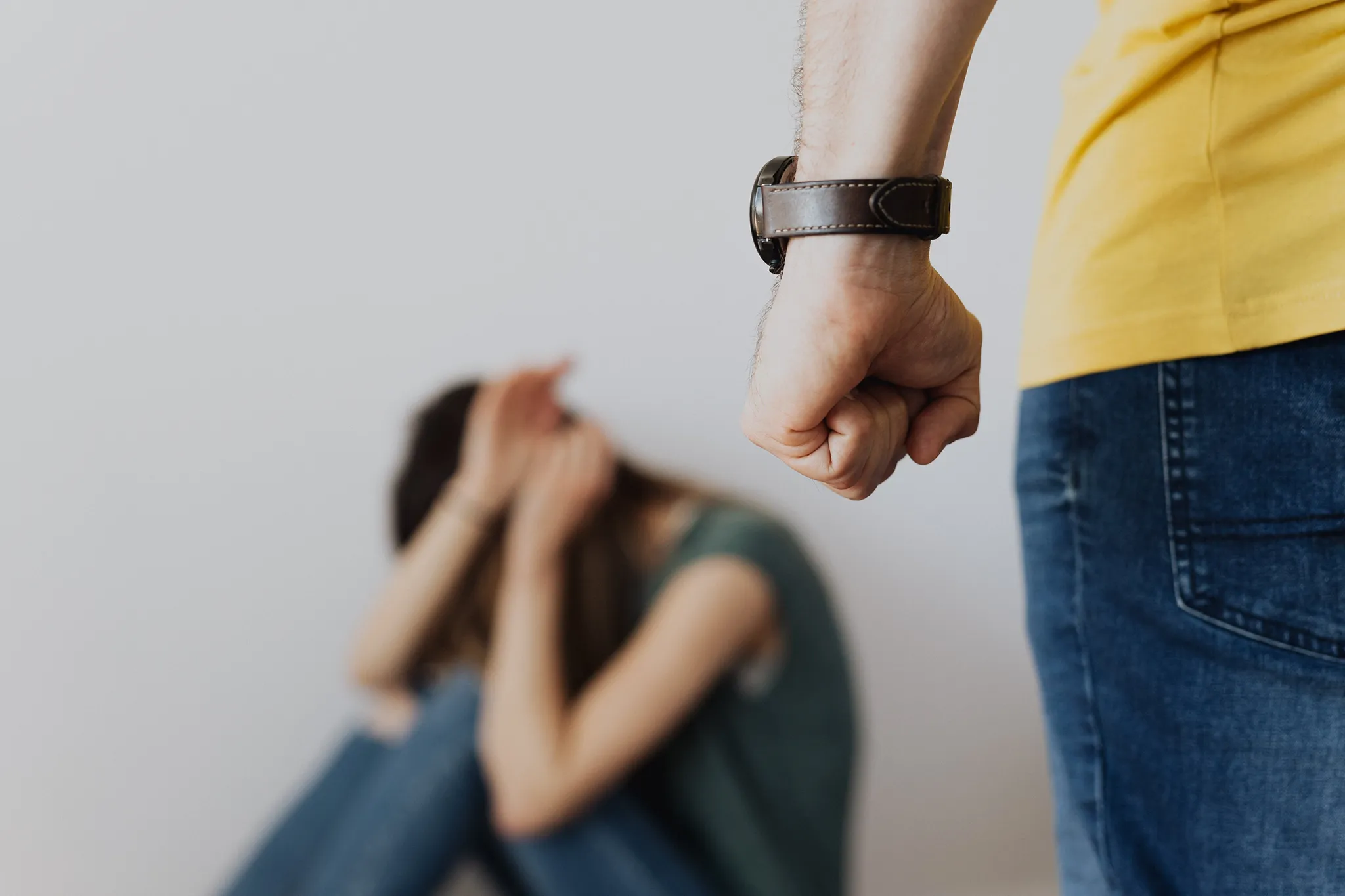 A man's fist is raised in the foreground, out of focus, while a blurred woman sits on the floor covering her face. This image depicts domestic violence or abuse.